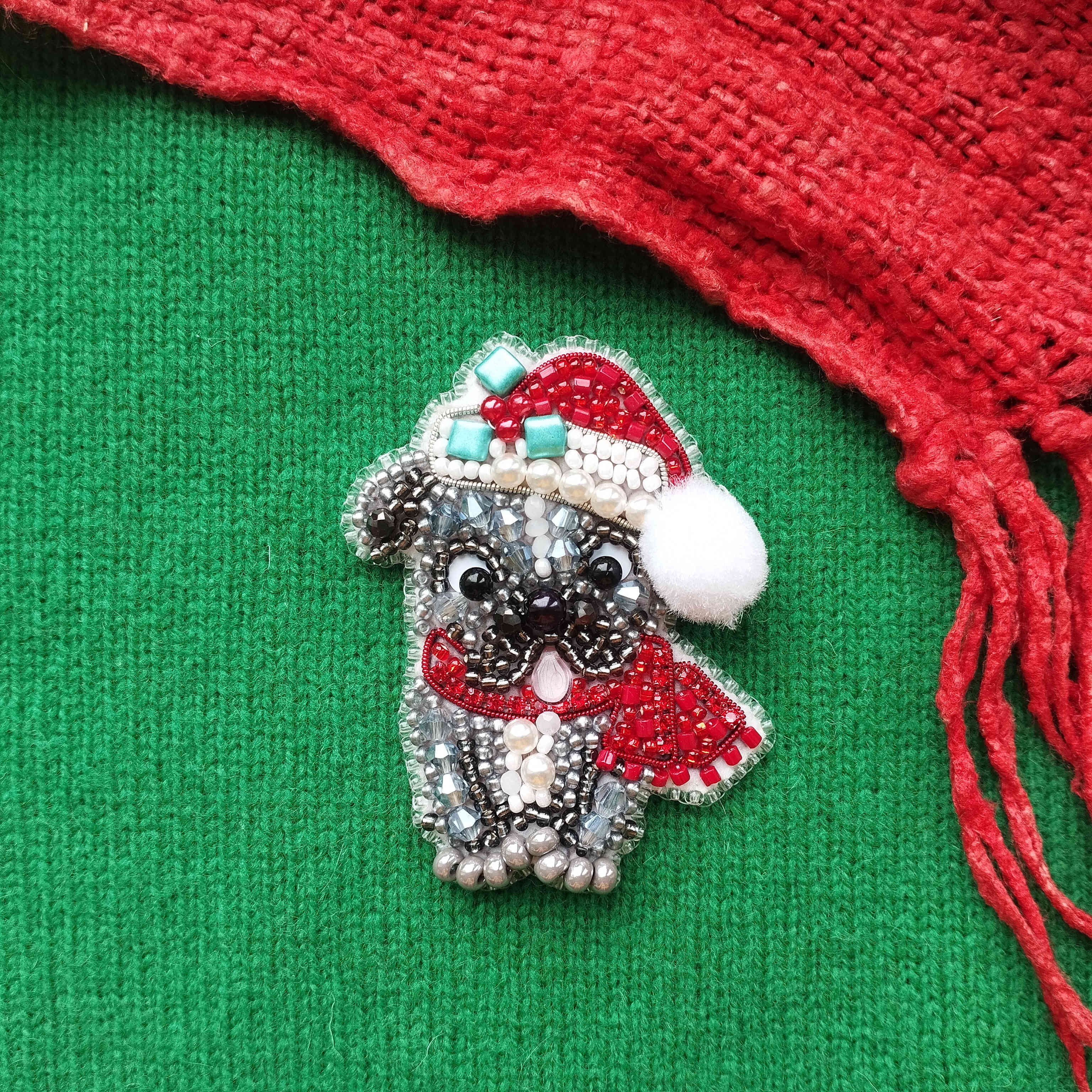 A decorative beaded brooch in the shape of a dog wearing a Santa hat and scarf, placed on a green fabric background with a red fringe.